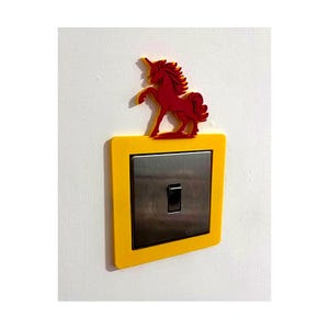 May include: A decorative light switch cover featuring a red unicorn silhouette atop a yellow frame. The switch plate has a silver finish with a black toggle switch. The cover is mounted on a white wall.