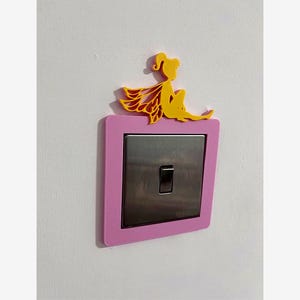 May include: A pink light switch cover with a yellow fairy design. The fairy is perched on top of the pink frame, which surrounds a silver switch plate. The switch is in the off position. The cover is mounted on a white wall.