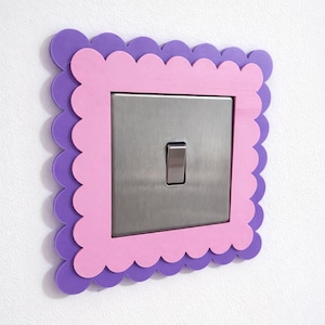 Peut inclure: Un cache-interrupteur décoratif avec un bord festonné. Le cadre est bicolore, avec un cadre intérieur rose et un cadre extérieur violet. L'interrupteur est en métal brossé.
