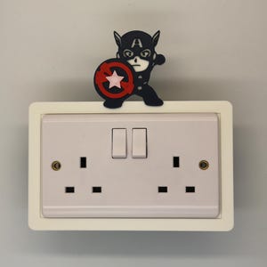 May include: A white electrical outlet with two sockets and two switches, framed in white. A black and blue Captain America figure with a shield is mounted on top, adding a decorative touch to the outlet.