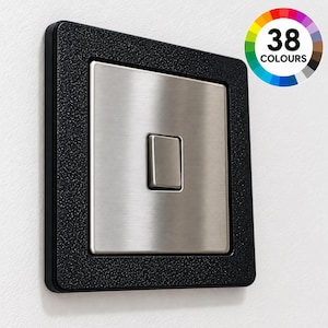 May include: A square light switch with a brushed silver metal finish and a central toggle, framed by a textured black border. A circular graphic in the top right corner displays a color wheel and the text 38 COLOURS, indicating variety.