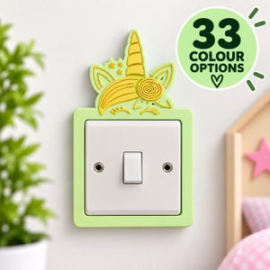 May include: A light green light switch cover featuring a yellow unicorn design with a horn, ears, and stars. The cover surrounds a standard white rectangular light switch. A circular graphic in the top right corner displays the text 33 COLOUR OPTIONS.