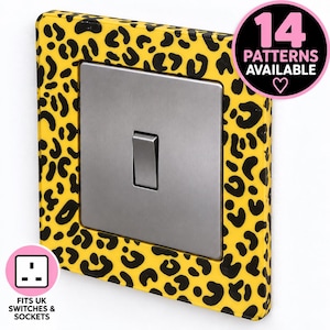 May include: A square light switch cover featuring a yellow background with a black leopard print pattern. The center contains a brushed silver metal toggle switch. A circular graphic in the corner states 14 PATTERNS AVAILABLE with a heart icon.