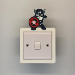 May include: A light switch with a white frame and a Captain America-themed decoration. The decoration features a cartoon figure holding a shield with a red star on a white background. The switch is centered on a light gray wall.