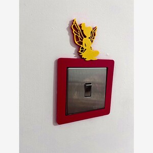 May include: A decorative light switch cover with a red frame and a silver switch. A yellow fairy-shaped accent sits above the switch. The switch is mounted on a white wall.