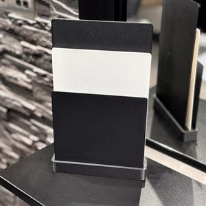 May include: A rectangular black and white sign holder featuring a clear base. The display consists of a tall black back panel, a white middle panel, and a shorter black front panel, all held upright by a transparent, thick rectangular base.