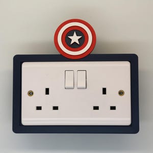 May include: A white double electrical socket with two toggle switches, framed in dark blue, with a red, white and blue Captain America shield on top.