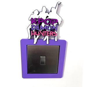 May include: A purple light switch cover with the text "KPOP DEMON HUNTERS" in white, purple, and pink. The cover has a silver switch and a white silhouette of three figures above the text.