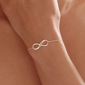Peut inclure: Un bracelet en argent avec un symbole infini et une chaîne délicate. Le bracelet est porté au poignet.