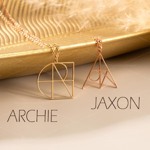 May include: Two gold-colored pendant necklaces. One pendant is a geometric design with the word "ARCHIE" below it. The other pendant is a triangle shape with the word "JAXON" below it. The necklaces are on a gold tray with a fluffy, beige background.