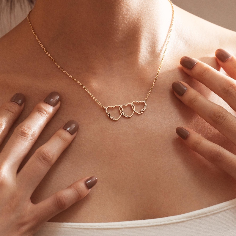 May include: A gold necklace with three interlocking heart charms. Each heart has a name inscribed inside: 'Katie', 'Liam', and 'Olivia'.