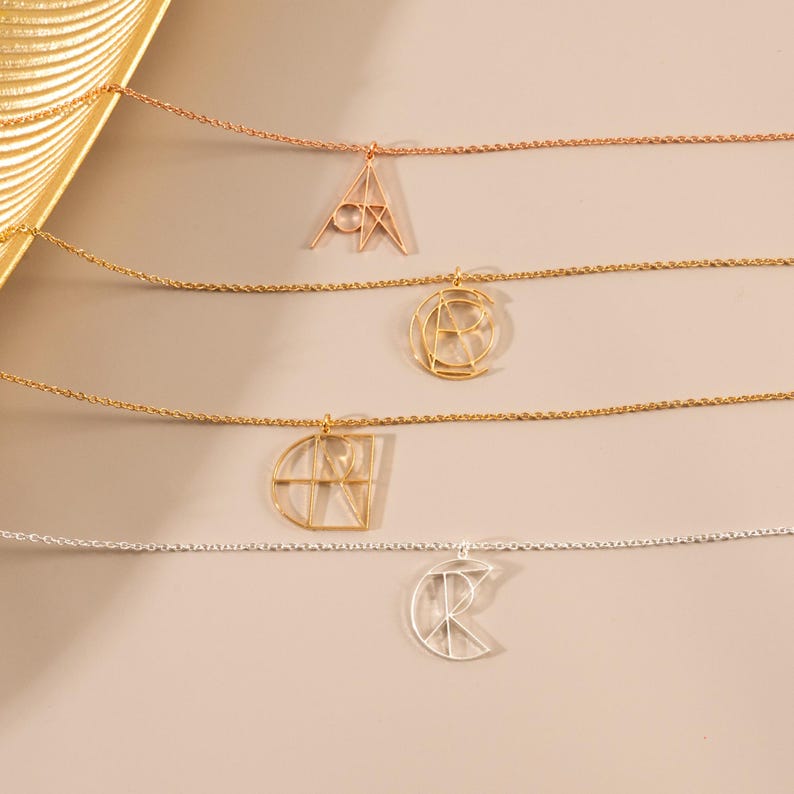 May include: Four gold necklaces with geometric letter pendants. The necklaces are layered on a beige surface. The pendants are in the shape of the letters A, C, R, and K.