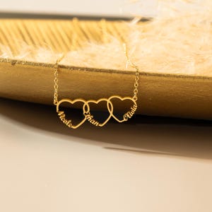K&ouml;nnte beinhalten: Goldkette mit drei ineinander verschlungenen Herzanh&auml;ngern, jeder mit einem eingravierten Namen: Nicole, Ethan und Olivia.
