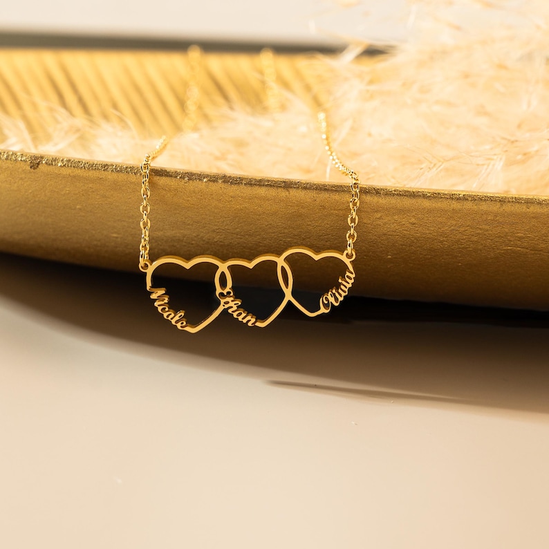 K&ouml;nnte beinhalten: Goldkette mit drei ineinander verschlungenen Herzanh&auml;ngern, jeder mit einem eingravierten Namen: Nicole, Ethan und Olivia.