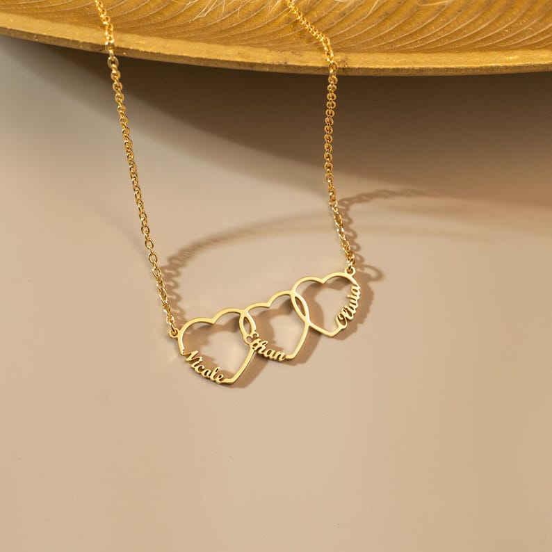 K&ouml;nnte beinhalten: Goldkette mit drei ineinandergreifenden Herzanh&auml;ngern, jeder mit einem eingravierten Namen: Nicole, Ethan und Olivia.
