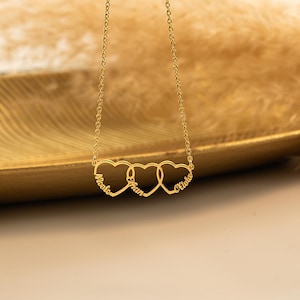 K&ouml;nnte beinhalten: Goldkette mit drei ineinander verschlungenen Herzanh&auml;ngern, jeder mit einem Namen graviert: "Nicole", "Ethan" und "Olivia".