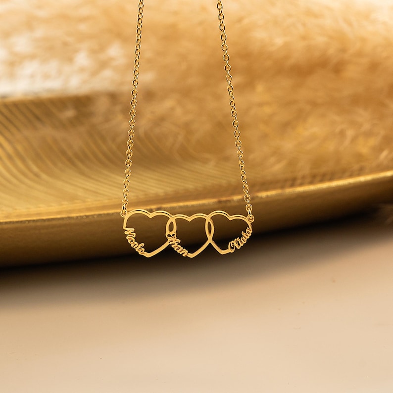 K&ouml;nnte beinhalten: Goldkette mit drei ineinander verschlungenen Herzanh&auml;ngern, jeder mit einem Namen graviert: "Nicole", "Ethan" und "Olivia".