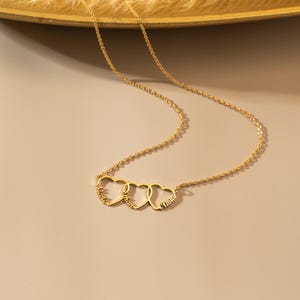K&ouml;nnte beinhalten: Goldkette mit drei ineinander verschlungenen Herzanh&auml;ngern, jeder mit einem Namen graviert: "Maria", "Ethan" und "Olivia".
