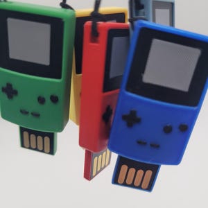May include: A collection of four handheld game console-shaped USB flash drives in green, yellow, red, and blue. Each device features a black screen area, a directional pad, two circular buttons, and a gold-colored USB connector at the base.