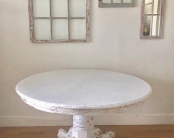 May include: White round table with a distressed finish and three legs shaped like animal paws. The table is in front of a white wall with three framed mirrors.