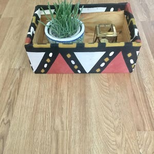 May include: A wooden storage box with a black, white, red, and yellow geometric pattern. The box contains a small potted plant and a gold candle holder.