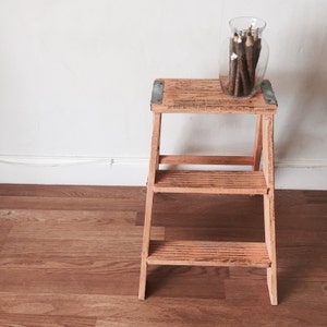 May include: A wooden step stool with two steps, painted in a light orange color. The stool has metal brackets on the top and bottom steps. A glass jar filled with colored pencils sits on the top step.