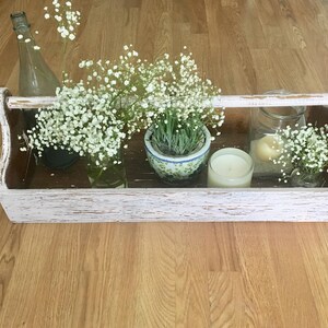May include: A whitewashed wooden tool box with a handle, filled with white baby's breath flowers, a clear glass bottle, a small potted plant, and two candles.