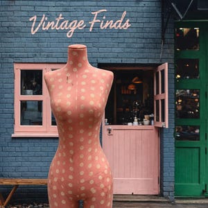 May include: A vintage-style dress form with a peach-colored fabric and cream polka dots stands in front of a blue brick building. The words "Vintage Finds" are written above the form. A pink door and green door are visible.