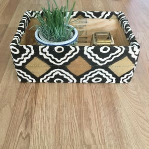 May include: A wooden storage box with a black, white, and gold geometric pattern. The box is filled with a small potted plant and a gold metal candle holder.