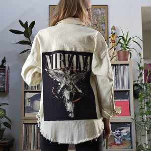 May include: A cream colored denim jacket with a black patch on the back that says "NIRVANA" with a graphic of an angel with wings.
