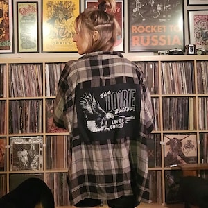 May include: A person wearing a black and white plaid flannel shirt with a black patch on the back that says "The Doobie Brothers Live in Concert".