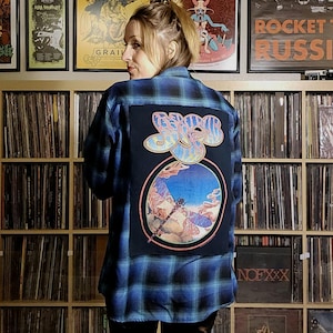May include: A blue and black plaid flannel shirt with a black patch on the back featuring a dragonfly and the text "Jethro Tull" in a colorful font.