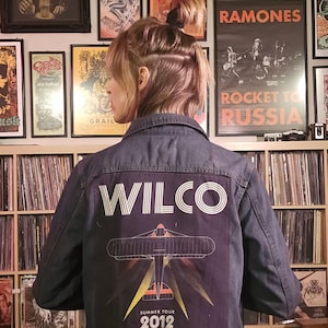 May include: A dark blue denim jacket with the band name "WILCO" printed in white across the back. Below the band name is a graphic of an airplane with the text "SUMMER TOUR 2012". The jacket has a button-up front.