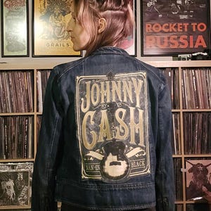 May include: A blue denim jacket with a gold and black Johnny Cash patch on the back. The patch features a guitar and the text "Johnny Cash", "Man in Black", and "American Recordings".