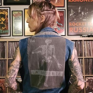 May include: A blue denim vest with a skeleton graphic on the back. The graphic is in black and white and says "14 LA MUERTE".