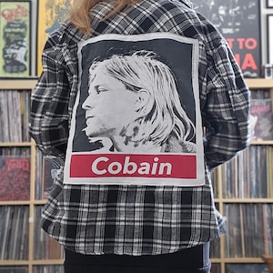 May include: A black and white plaid flannel shirt with a large patch featuring a portrait of Kurt Cobain and the name "Cobain" in a red box. The person is wearing a gray beanie and black skirt.