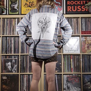 May include: A blue and gray striped button-down shirt with a gray graphic on the back. The graphic depicts a figure on a motorcycle. The person is wearing a patterned skirt and black boots. The background features a wall of vinyl records and concert posters.