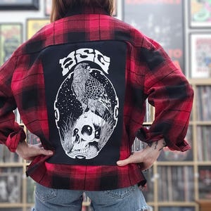 May include: A red and black plaid flannel shirt with a black patch on the back. The patch features a black and white illustration of a bird perched on a skull with the word "BIG" written in a stylized font.