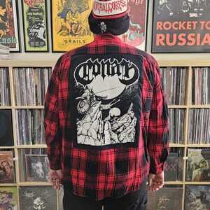 May include: A red and black plaid flannel shirt with a black patch on the back. The patch features a black and white illustration of a creature with a large head and horns. The text "Opprobrium" is printed in white on the patch.