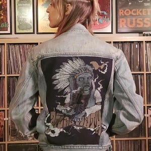 May include: A light blue denim jacket with a black patch on the back. The patch features a drawing of a Native American person wearing a headdress and holding a pipe. The text "Peace Out" is written below the image.