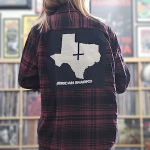 May include: A person wearing a red and black plaid flannel shirt with a black patch on the back. The patch features a white outline of the state of Texas with a cross inside and the text "American Sharks" below.