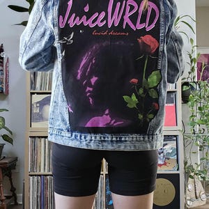 May include: A light blue denim jacket with a black patch on the back. The patch features a pink and white graphic of the text "Juice WRLD" with the words "lucid dreams" underneath. A silhouette of a person with a rose is in the background.