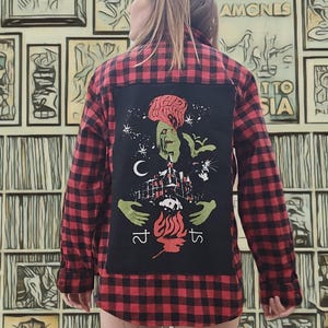 May include: Red and black plaid flannel shirt with a black graphic patch on the back. The patch features a green zombie-like figure, a building, and the text "215". The shirt is worn over bare legs.