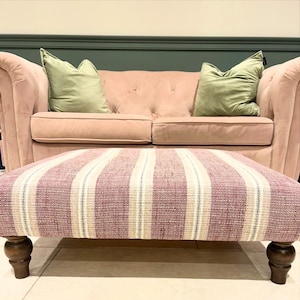 May include: A pink velvet loveseat with two sage green throw pillows. In front of the loveseat is a rectangular ottoman with a striped pattern in red, white, and beige. The ottoman has dark wooden legs.