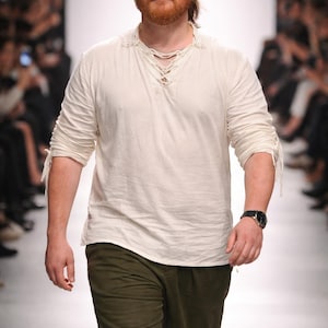May include: A person modeling a cream-colored long-sleeve shirt with a laced neckline and olive green pants. The model is wearing a black watch with a silver-colored face. The background is a fashion show runway.