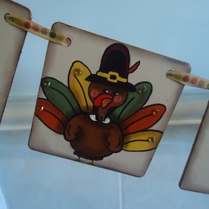 May include: A Thanksgiving banner with a painted turkey wearing a pilgrim hat. The turkey is brown with red, yellow, green, and orange feathers. The banner is made of wood and has a brown background.