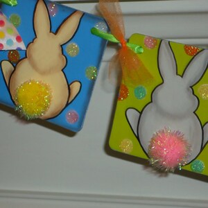 May include: A banner with two painted bunnies with fluffy tails. The bunny on the left is yellow and the bunny on the right is white. The banner is decorated with colorful polka dots.