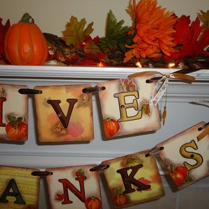 May include: A Thanksgiving banner with the word "GIVENS" spelled out in brown letters on a beige background with fall leaves and small pumpkins. The banner is strung with brown ribbon and is hanging on a white mantel.