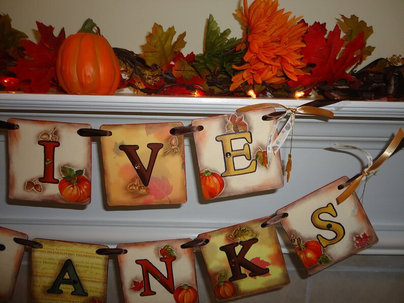 May include: A Thanksgiving banner with the word "GIVENS" spelled out in brown letters on a beige background with fall leaves and small pumpkins. The banner is strung with brown ribbon and is hanging on a white mantel.