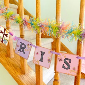 May include: A purple, pink, and gold banner with the words "He Is Risen" hanging on a wooden staircase railing. The banner is decorated with flowers, butterflies, and glitter.